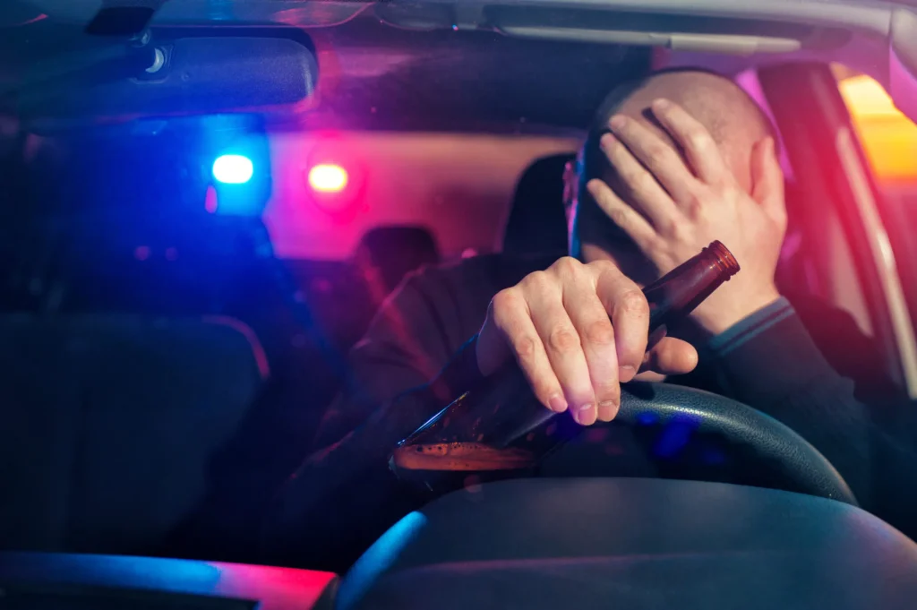 Man sitting in a car holding a beer bottle, covering his face, with police lights flashing in the background, illustrating the emotional impact of a DUI arrest in Texas.