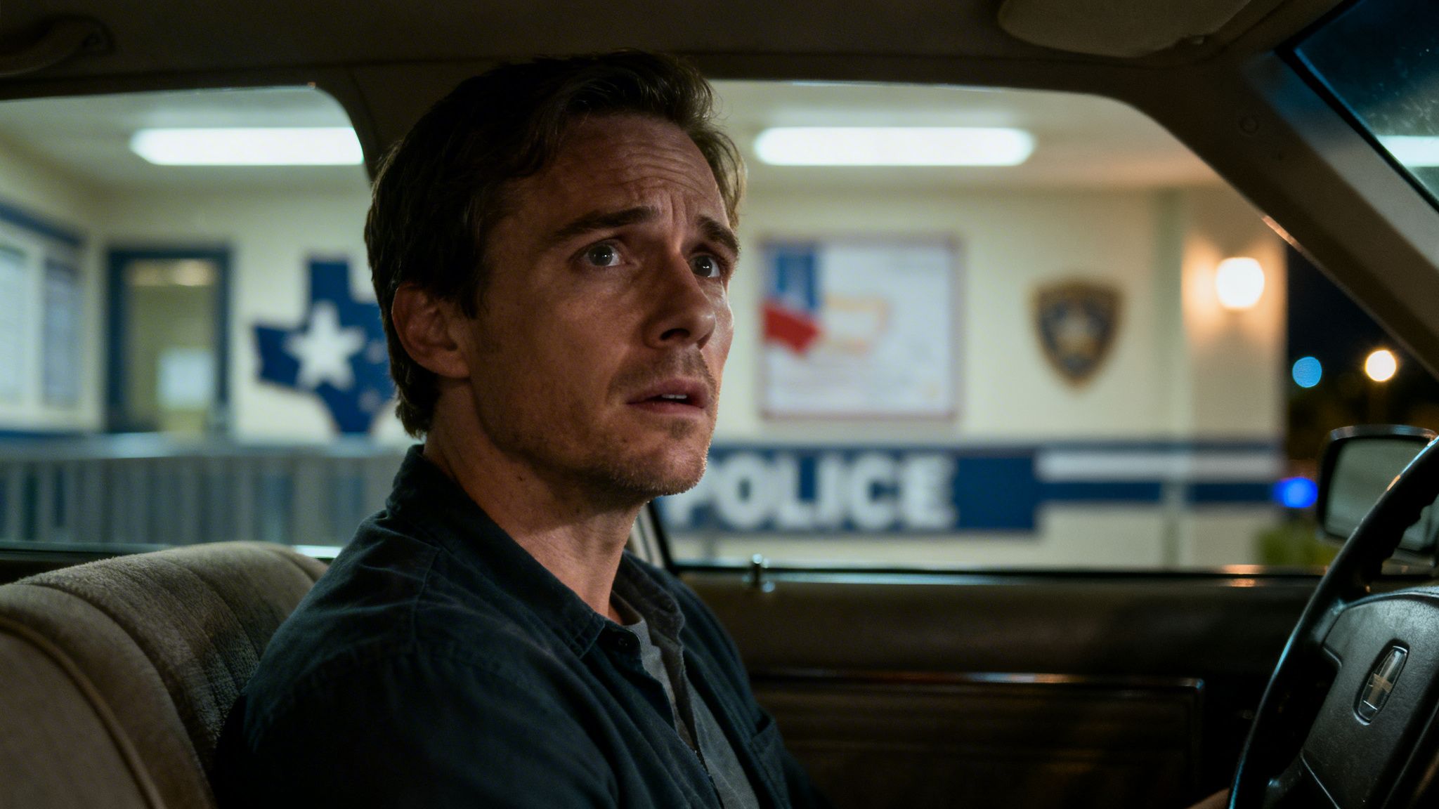 A concerned man sitting in a car, looking towards a Texas police station in the background.