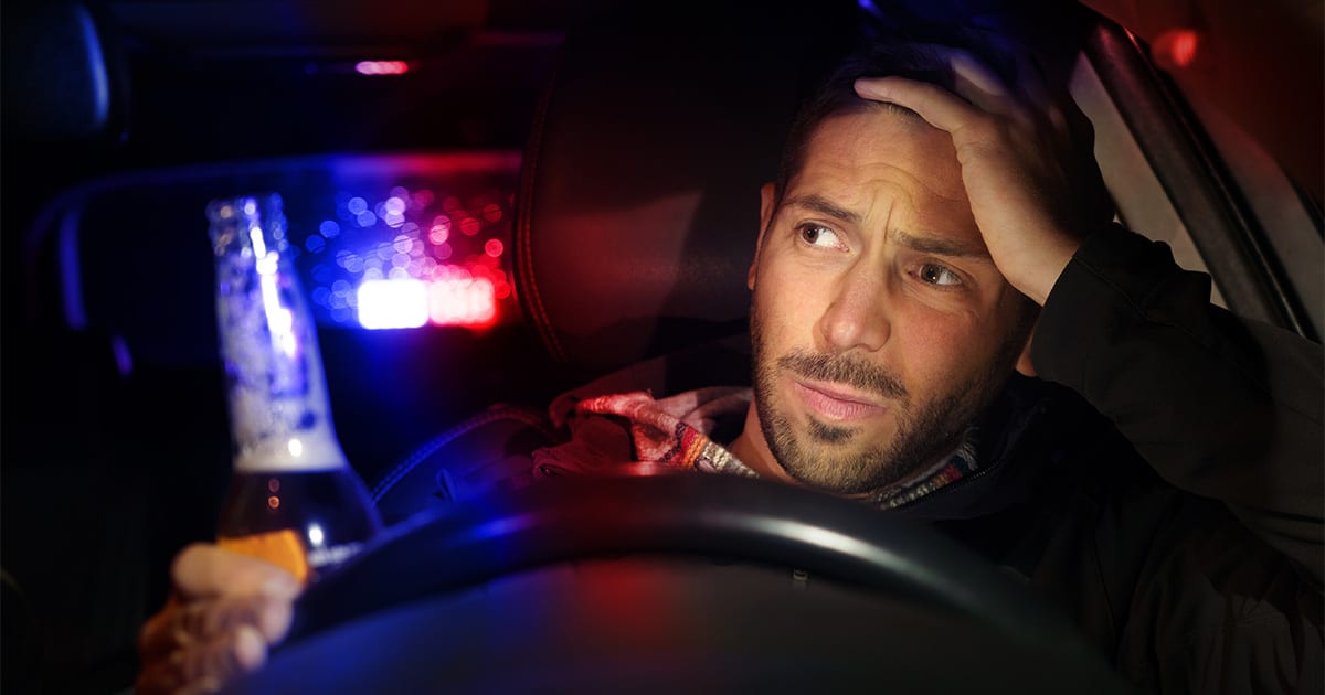 Man in car holding a beer bottle, looking worried with police lights flashing in the background, illustrating the consequences of a DWI arrest.