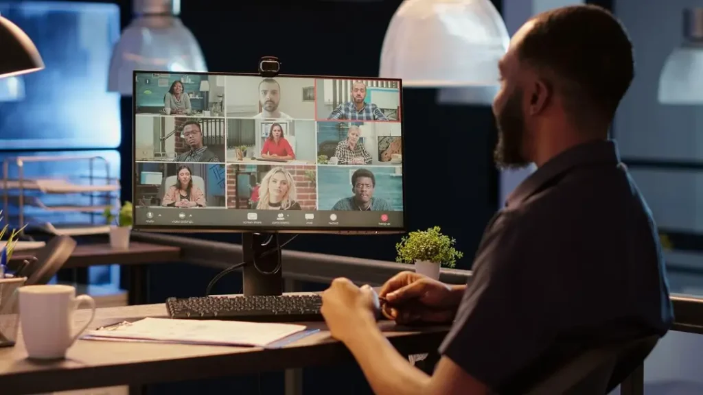 Man participating in a video consultation on a computer screen, featuring multiple attendees, in a modern office setting with a coffee cup and documents on the desk.