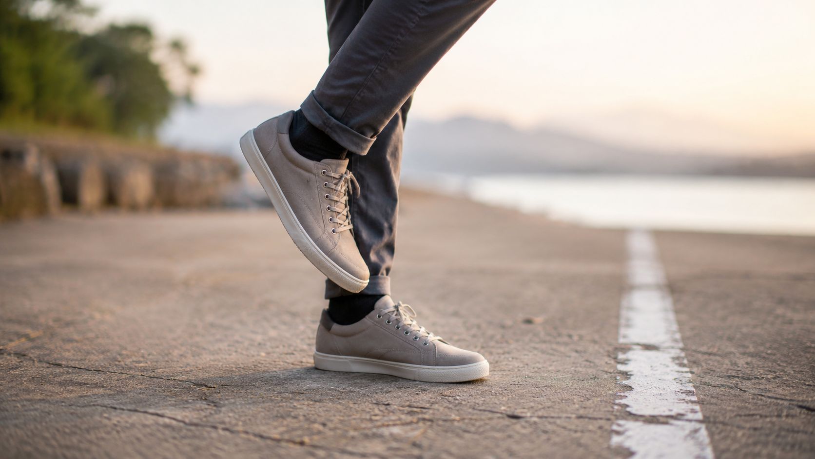 A person standing on a paved road wearing casual grey sneakers and rolled-up trousers at sunset.